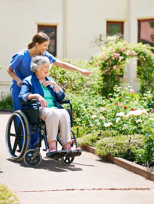 Eine Pflegerin schiebt eine ältere Frau im Rollstuhl durch einen blühenden Garten mit rosafarbenen und weißen Rosen. Die Pflegerin zeigt auf etwas in der Umgebung, während die ältere Dame interessiert schaut. Der Weg ist gepflastert und von Pflanzen gesäumt.