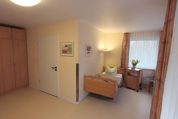Der Raum ist hell und freundlich gestaltet. Auf der linken Seite steht ein Bett mit einem Kissen, auf der rechten Seite befindet sich ein Tisch mit frischen Blumen. Eine große Fensterfront sorgt für Tageslicht, und ein Kleiderschrank ergänzt die Einrichtung.