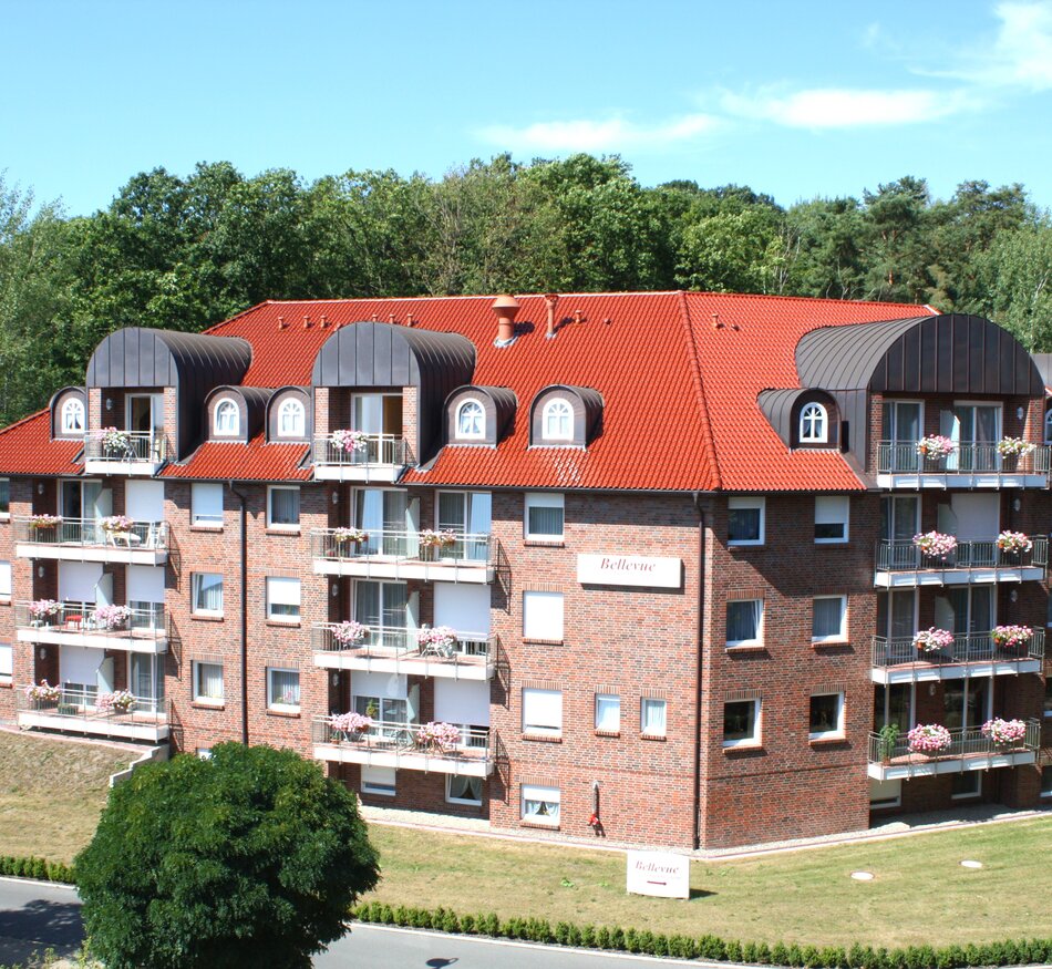 Ein mehrstöckiges, rotes Ziegelgebäude mit einem gewölbten Dach und Balkonen ist umgeben von Bäumen und Rasenflächen. Das Gebäude hat mehrere Fenster und eine freundliche Erscheinung. Es deutet darauf hin, dass es sich um Wohnraum handelt.