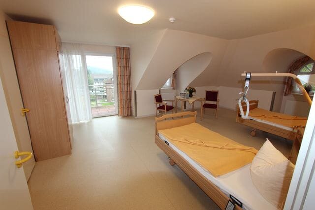 Ein helles Zimmer mit zwei Pflegebetten, einem Tisch und Stühlen. Eine Tür führt zu einem Balkon mit Aussicht. An einem Bett befindet sich ein Haltegriff zur Unterstützung. Die Wände sind in neutralen Farben gehalten, und das Licht ist weich.