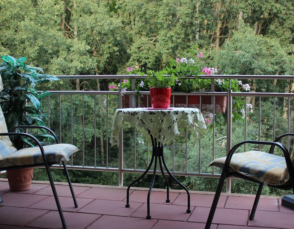 Der Balkon ist mit zwei Stühlen und einem kleinen Tisch ausgestattet. Auf dem Tisch steht ein Blumentopf mit einer grünen Pflanze. Im Hintergrund sind Bäume und Blumen zu sehen, die dem Ort eine ruhige und einladende Atmosphäre verleihen.