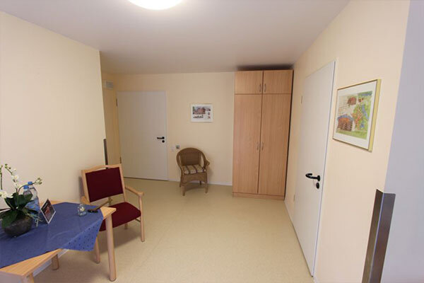 Ein heller Raum mit einem hellen Boden und beige Wänden. Auf der linken Seite steht ein Tisch mit einer blauen Tischdecke und einer Pflanze. Daneben steht ein roter Stuhl. Gegenüber befindet sich ein Kleiderschrank und eine geschlossene Tür. An der Wand hängt ein Bild.