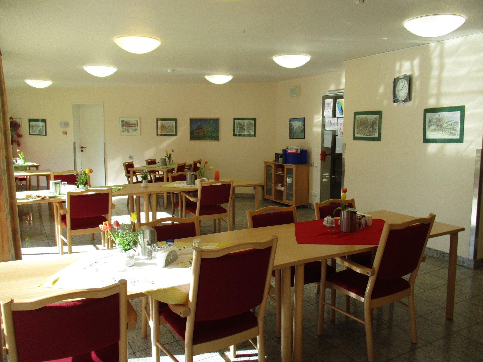 Ein helles und freundliches Esszimmer mit mehreren Tischen und rot gepolsterten Stühlen. Auf den Tischen stehen Blumenvasen und Geschirr. An den Wänden hängen Bilder. Das Ambiente ist einladend und gemütlich, ideal für gesellige Treffen.