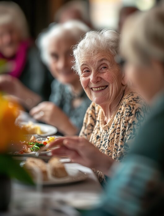 Eine Gruppe älterer Frauen sitzt an einem Tisch und genießt eine Mahlzeit. Eine Frau in der Mitte mit grauen Haaren lächelt fröhlich in die Kamera. Blumenarrangements sind im Hintergrund zu sehen, was eine festliche Atmosphäre schafft.