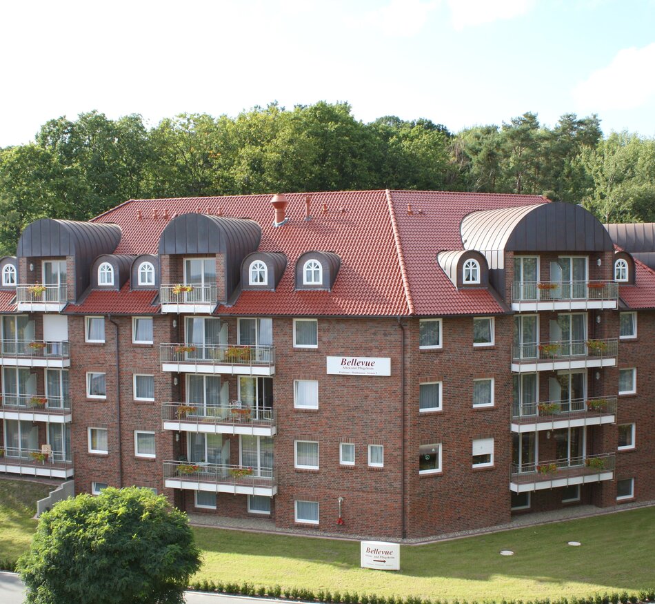 Das Bild zeigt ein mehrstöckiges, reddish-braunes Wohngebäude mit Balkon. Das Dach hat abgerundete Gauben. Vor dem Gebäude befinden sich Bäume und eine gepflegte Rasenfläche. Es wirkt modern und einladend.
