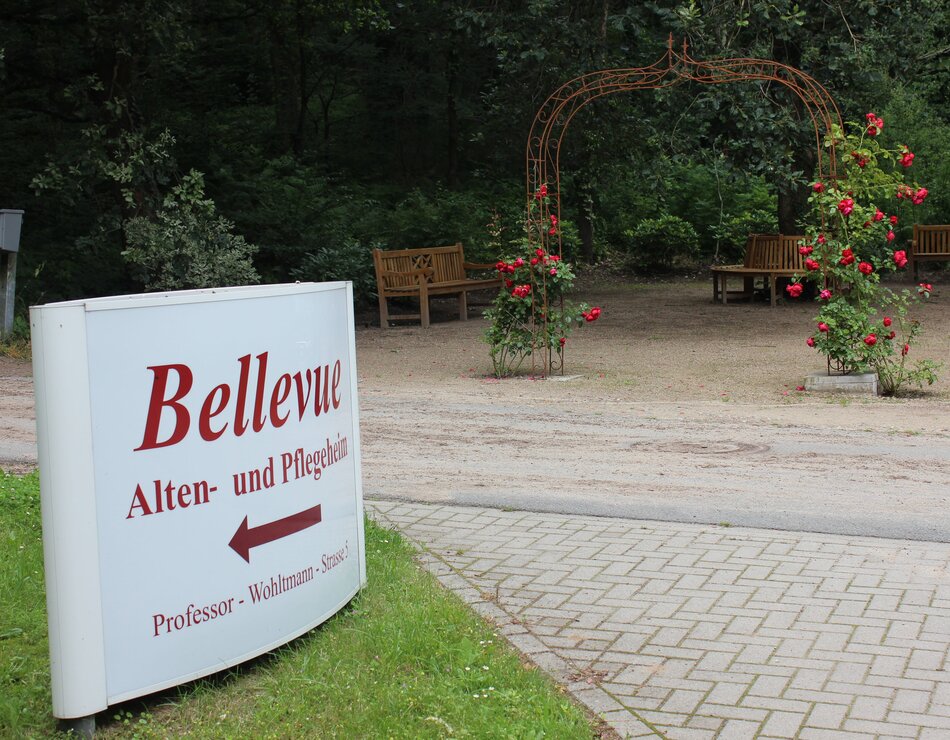 Ein Schild mit der Aufschrift „Bellevue Alten- und Pflegeheim“ zeigt nach links. Im Hintergrund ist ein Weg mit Bänken und einem Rosenbogen zu sehen, umgeben von Bäumen. Dieses Bild vermittelt den Eindruck eines einladenden und ruhigen Pflegebereichs.