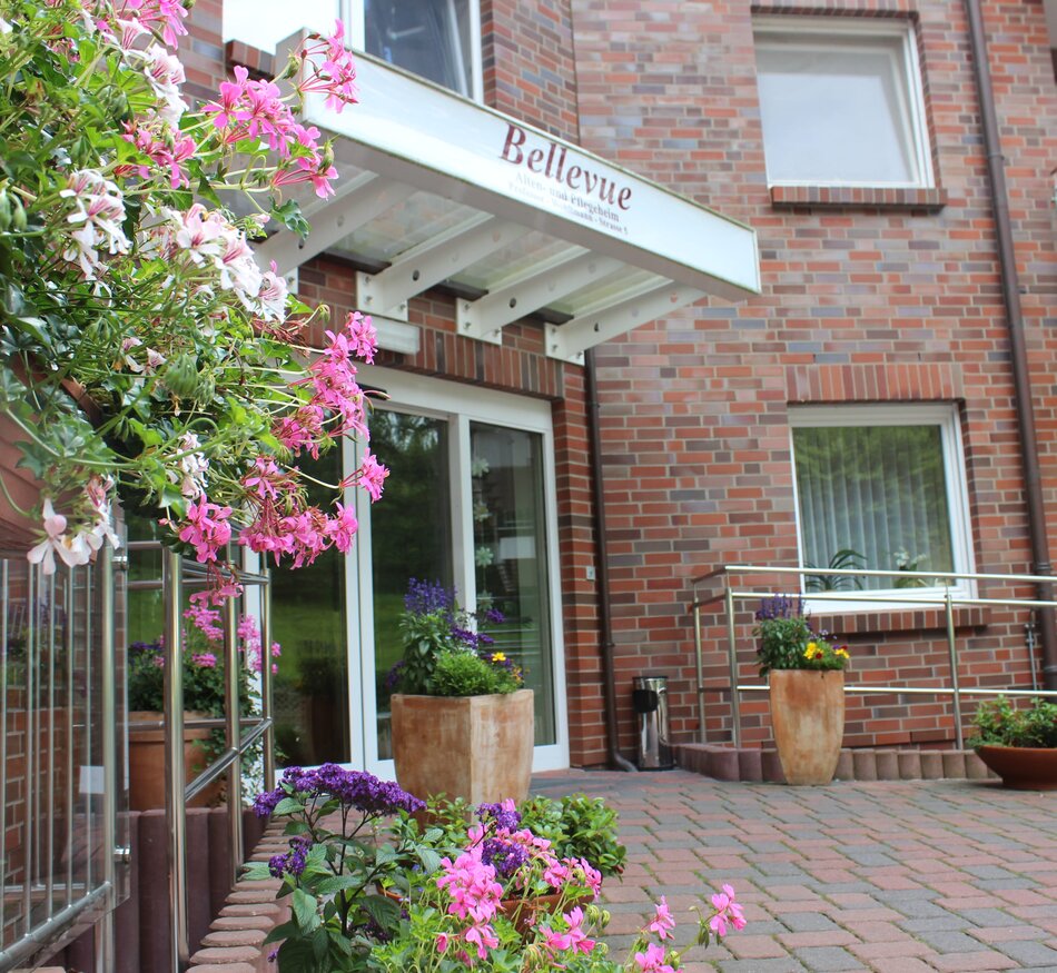 Ein blühender Eingangsbereich zeigt einen gepflasterten Weg vor einem Gebäude mit einem Schild "Bellevue". Blühende Pflanzen in Töpfen und hängende Blumenampeln flanken den Eingang. Die Atmosphäre wirkt einladend und freundlich.