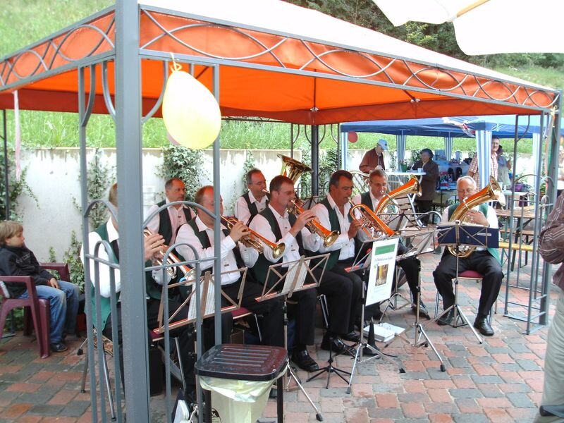 Eine Blaskapelle spielt unter einem orangefarbenen Pavillon. Die Musiker tragen festliche Kleidung und spielen verschiedene Blasinstrumente. Im Hintergrund sind weitere Stände und eine grüne Wiese sichtbar. Die Szenerie vermittelt eine fröhliche, festliche Atmosphäre.