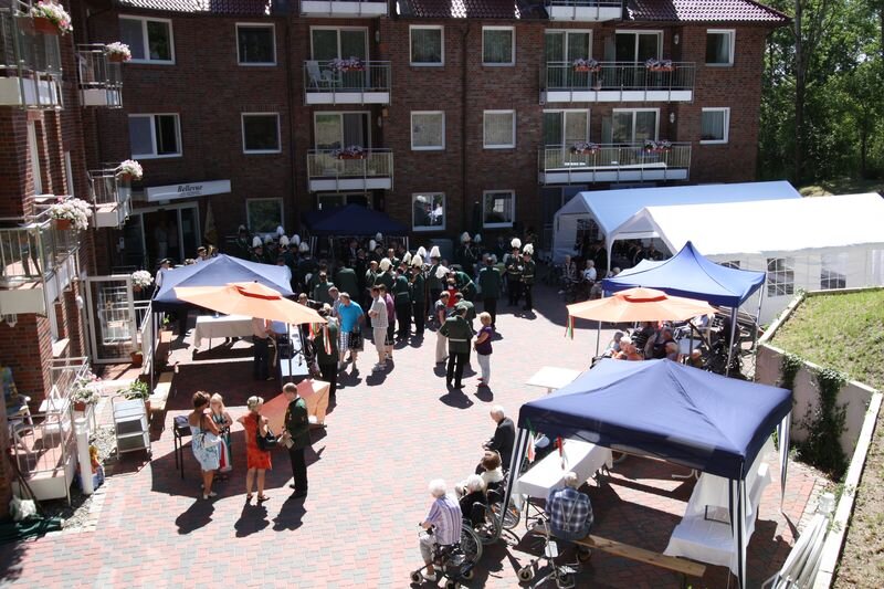 Eine Menschenmenge versammelt sich in einem Innenhof, umgeben von Wohngebäuden. Es gibt verschiedene Pavillons und Stände. Einige Personen stehen im Mittelpunkt des Geschehens, während andere in der Nähe sitzen oder stehen. Es herrscht eine festliche Atmosphäre.