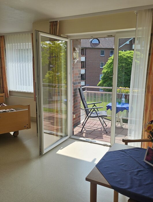 Ein helles Zimmer mit einem Bett, einem Tisch und Stühlen. Große Fenster führen zu einem Balkon mit Gartenblick. Auf dem Tisch steht eine Vase mit Blumen. Die Einrichtung wirkt freundlich und einladend.