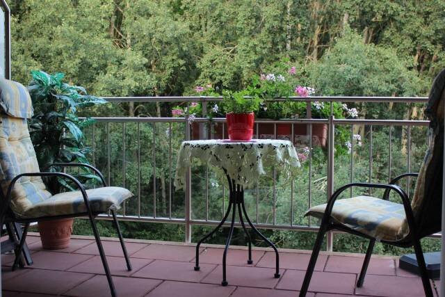 Ein Balkon mit zwei Stühlen und einem runden Tisch in der Mitte. Auf dem Tisch steht ein Blumentopf mit einer Pflanze. Im Hintergrund sind Bäume und Blumen zu sehen, die eine gemütliche, grüne Umgebung schaffen.