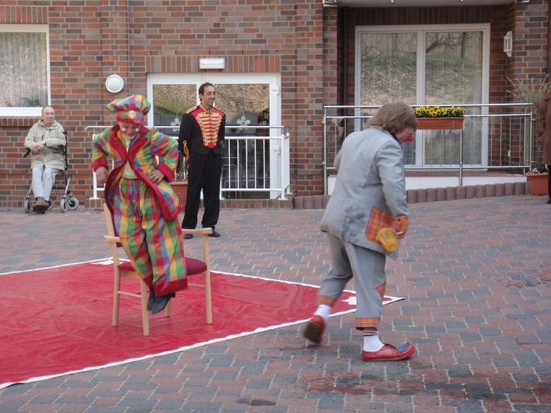Ein Clown in bunter Kostümierung balanciert auf einem Stuhl, während ein weiterer Clown in grauer Kleidung mit einer Kiste auf dem Weg zum Zentrum des Geschehens ist. Im Hintergrund sitzen Zuschauer, die die Aufführung beobachten.