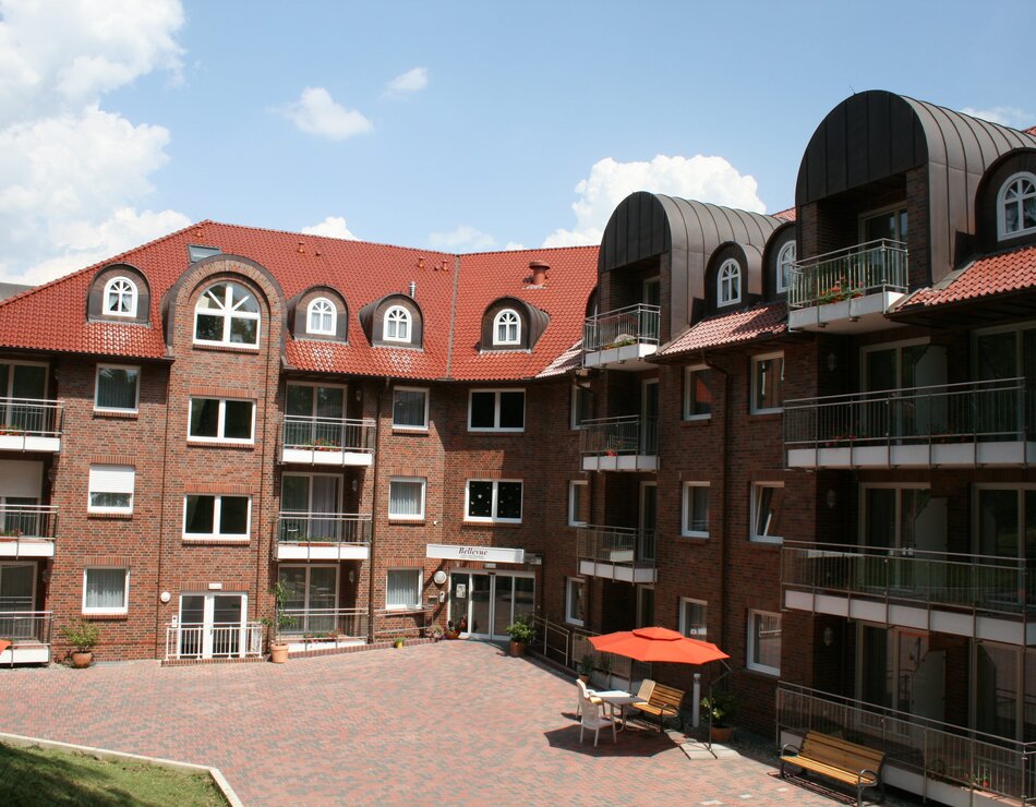 Pflegeheim mit rot gedeckten Dach und mehreren Balkonen. Der Innenhof ist gepflastert, mit zwei roten Sonnenschirmen und Sitzgelegenheiten. Der Himmel ist blau mit einigen Wolken.