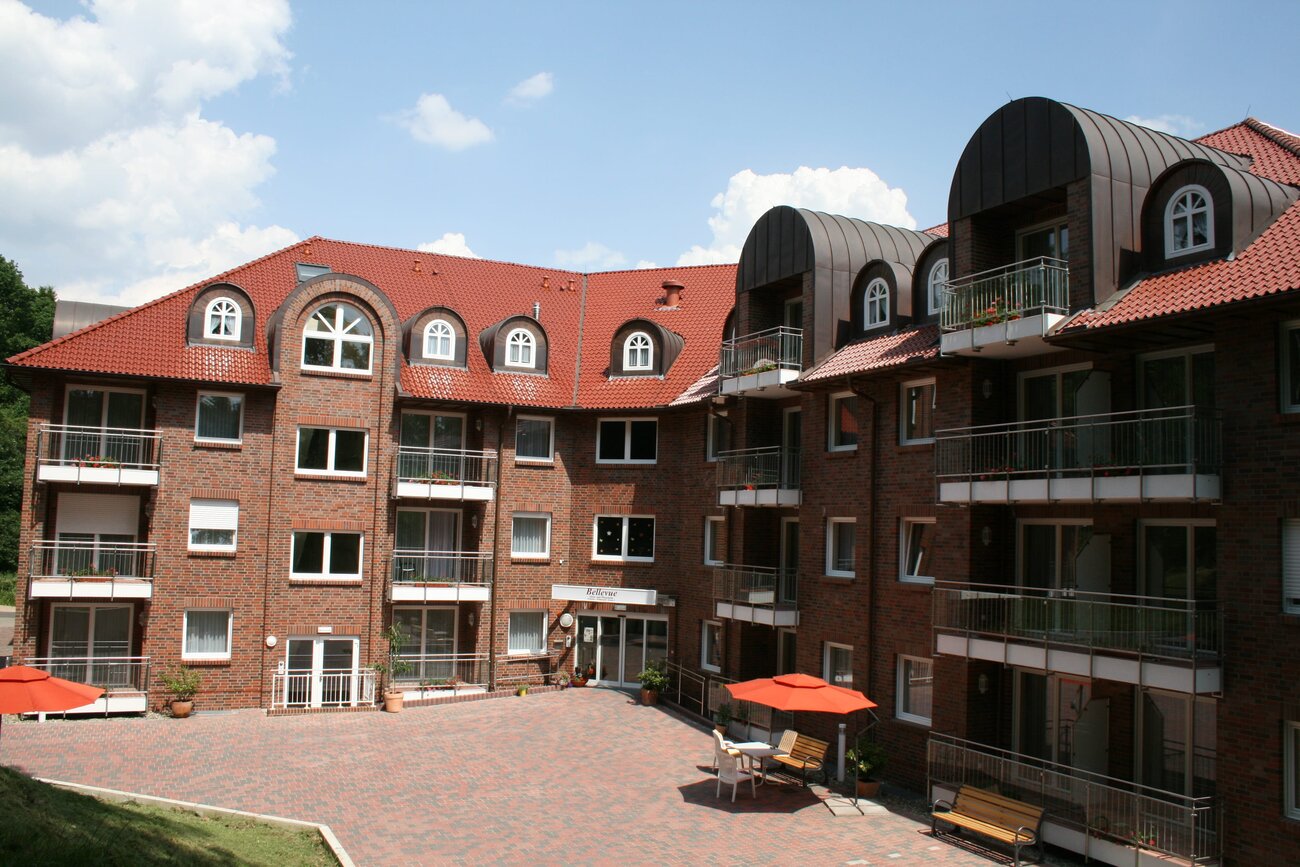Pflegeheim mit rot gedeckten Dach und mehreren Balkonen. Der Innenhof ist gepflastert, mit zwei roten Sonnenschirmen und Sitzgelegenheiten. Der Himmel ist blau mit einigen Wolken.