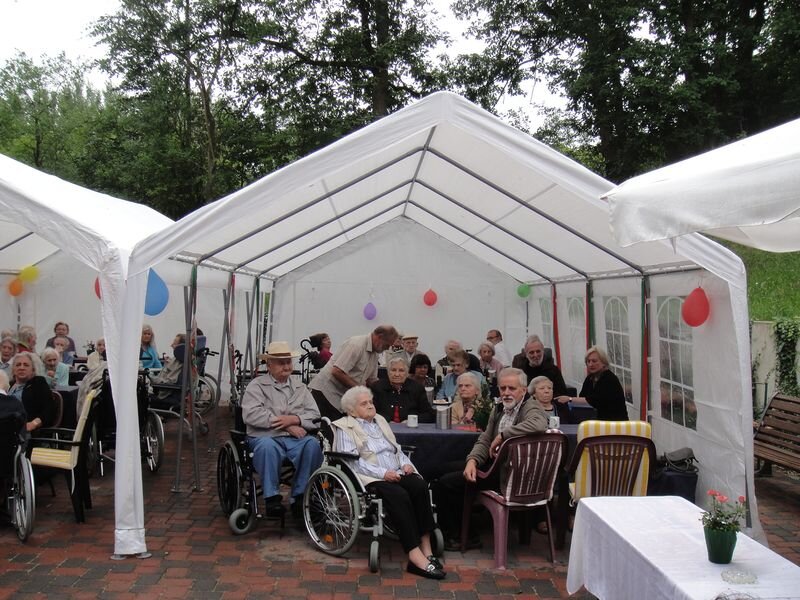 Eine Gruppe älterer Menschen sitzt in einem Zelt im Freien, umgeben von Tischen und Stühlen. Einige Personen sind im Rollstuhl, andere stehen oder sitzen. Ballons schmücken das Zelt, und es scheint eine gesellige Atmosphäre zu herrschen.