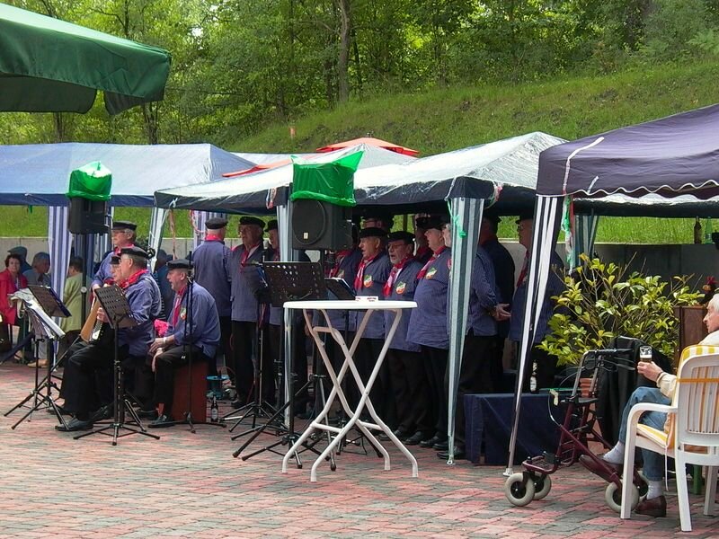 Eine Gruppe von Sängern in blauer Kleidung steht unter Zelten und singt. Einige Musiker spielen Instrumente. Im Hintergrund sind grüne Bäume sichtbar. Die Atmosphäre wirkt festlich und gesellig.