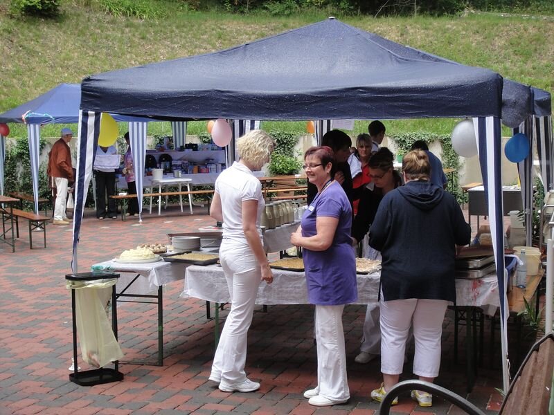 Eine Gruppen von Personen steht unter einem Festzelt, während sie Essen und Getränke servieren. Einige Gäste stehen in der Nähe der Tische, die für ein gemeinsames Festmahl vorbereitet sind. Es herrscht eine gefällige, gesellige Atmosphäre.
