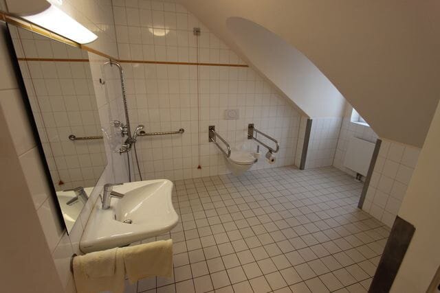 Behindertenfreundliches Badezimmer mit einem Waschbecken, einem Handtuch, einer Duschkabine und einem Toilettenbereich. Die Wände sind gefliest, und das Badezimmer verfügt über Haltestangen für zusätzliche Unterstützung. Das Dach ist schräg.