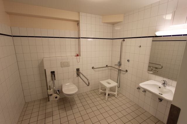 Das Bild zeigt ein barrierefreies Badezimmer. Es enthält eine Toilettenschüssel mit Haltegriffen, eine Duschbank und einen Waschplatz mit Spiegel. Die Wände sind gefliest und der Raum wirkt hell und zugänglich.