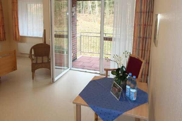 Ein heller Raum mit Zugang zu einem Balkon. In der Mitte steht ein Tisch mit einer blauen Tischdecke, darauf eine Blumenvase und zwei Flaschen Wasser. Neben dem Tisch befindet sich ein Stuhl und ein rattanartiger Sessel. Fenster lassen Tageslicht herein.