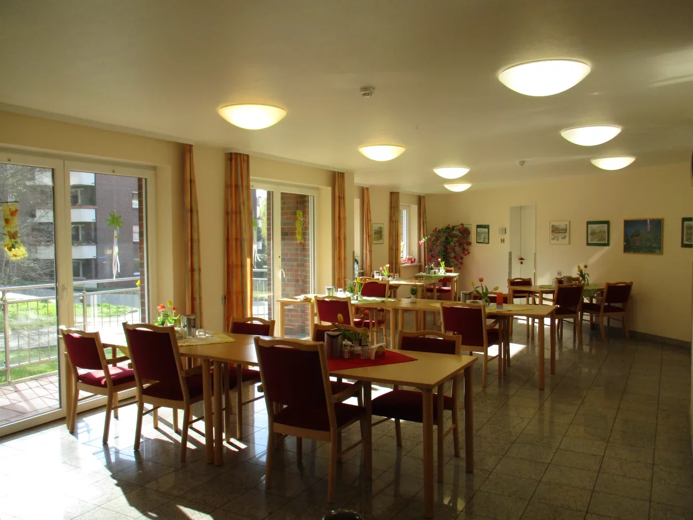 Ein heller Speisesaal mit mehreren Tischen und roten Stühlen. Große Fenster lassen Tageslicht herein und bieten Blick ins Freie. Einige Tische sind dekoriert, und Pflanzen sind in der Ecke zu sehen. Die Atmosphäre wirkt einladend und freundlich.