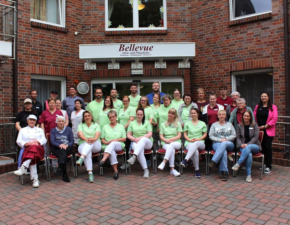 Eine große Gruppe von Personen posiert vor einem Gebäude mit dem Schild "Bellevue". Die meisten tragen grüne T-Shirts. Einige Personen sitzen, während andere stehen. Es herrscht eine freundliche Atmosphäre, die Gemeinschaft und Teamarbeit vermittelt.