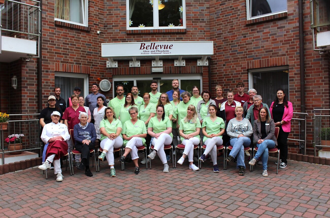 Eine große Gruppe von Personen posiert vor einem Gebäude mit dem Schild "Bellevue". Die meisten tragen grüne T-Shirts. Einige Personen sitzen, während andere stehen. Es herrscht eine freundliche Atmosphäre, die Gemeinschaft und Teamarbeit vermittelt.