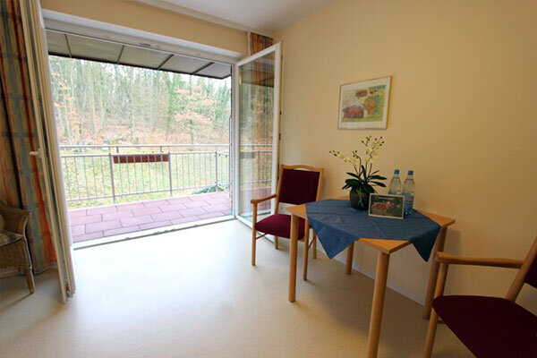Der Raum zeigt einen kleinen Tisch mit zwei Stühlen und einer Pflanze sowie zwei Wasserflaschen. Ein großes Fenster öffnet sich zu einem Balkon mit Blick auf einen bewaldeten Bereich. Der Fußboden ist hell und der Raum wirkt freundlich und einladend.