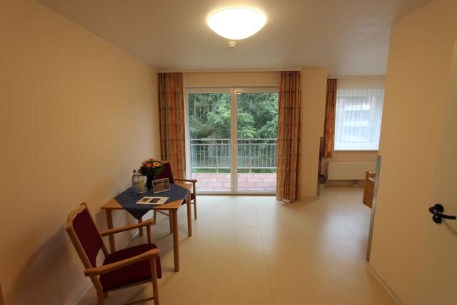 Ein heller Raum mit einem Tisch und zwei Stühlen. Auf dem Tisch stehen Wasserflaschen und eine Blumenvase. Große Fenster führen zu einem Balkon mit Blick ins Grüne. Die Wände sind in neutralen Farben gehalten, und der Boden ist hell.