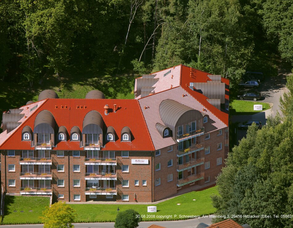 Das Bild zeigt ein mehrstöckiges Wohngebäude mit roten Dachziegeln und Balkonen. Umgeben von Bäumen, steht das Gebäude in einer ländlichen Umgebung und hat eine gelbe Fassade. Es vermittelt einen Eindruck von Ruhe und Naturverbundenheit.