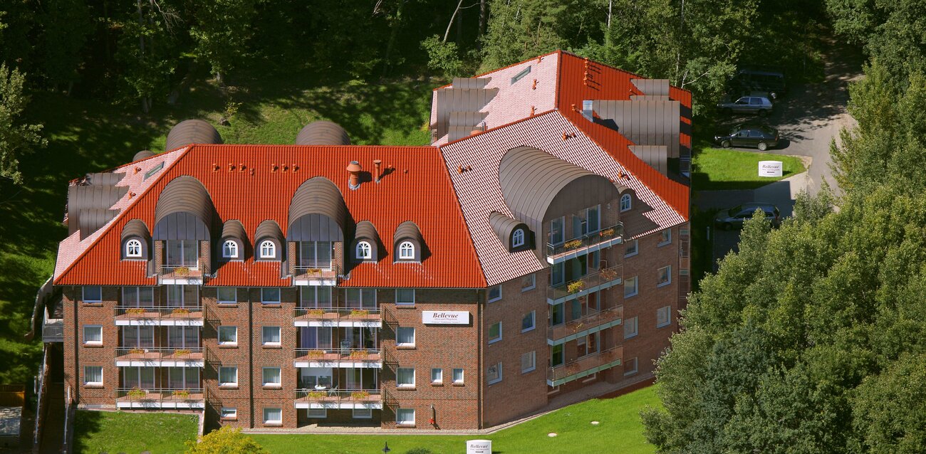 Das Bild zeigt ein mehrstöckiges Wohngebäude mit roten Dachziegeln und Balkonen. Umgeben von Bäumen, steht das Gebäude in einer ländlichen Umgebung und hat eine gelbe Fassade. Es vermittelt einen Eindruck von Ruhe und Naturverbundenheit.