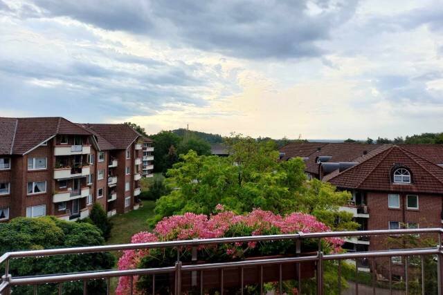 Ein Blick von einem Balkon auf eine grüne Umgebung mit blühenden Pflanzen im Vordergrund. Im Hintergrund sind mehrere Wohngebäude zu sehen, unter einem bewölkten Himmel. Die Szene vermittelt eine ruhige, reizvolle Atmosphäre.