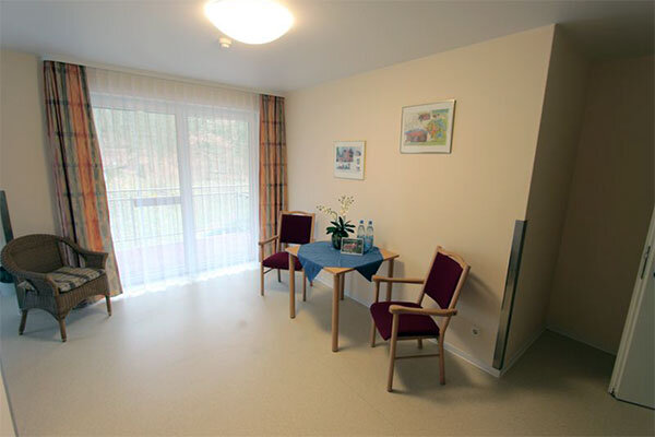 Ein heller Raum mit einem Tisch und zwei Stühlen. Auf dem Tisch stehen Wasserflaschen und eine Pflanze. An der Wand hängen zwei Bilder. Durch große Fenster strömt Tageslicht in den Raum, der insgesamt einladend und freundlich wirkt.